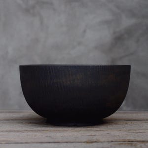 以下が含まれることがあります： マット仕上げの黒い丸い木製のボウル。ボウルは木製の表面に置かれています。ボウルの内側は見えません。背景はニュートラルグレーです。