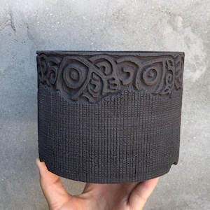 May include: A dark grey ceramic planter with a textured surface. The top edge features a carved, repeating swirl design. The lower portion has a vertical, lined texture. The planter is held in a hand.