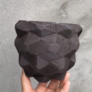 Könnte beinhalten: Ein dunkelbrauner Keramik-Pflanztopf mit geometrischem Muster. Der Pflanztopf hat eine strukturierte Oberfläche mit geschnitzten Linien, die einen 3D-Effekt erzeugen. Der Topf wird in einer Hand vor grauem Hintergrund gehalten.