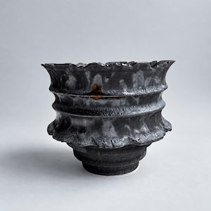 Puede incluir: Un cuenco de cerámica gris carbón oscuro con un borde ancho, acampanado y dentado. El exterior presenta una serie de crestas horizontales y un acabado mate texturizado con sutiles reflejos metálicos y una mancha de oxidación rojiza.