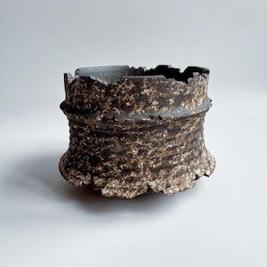 May include: A white ceramic mug with a black handle and the text "But First, Coffee" in a bold, black font. The mug is set against a blurred background. The mug appears to be approximately 10 cm tall.