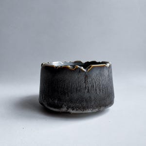 以下が含まれることがあります： 暗いチャコールグレーの外観を持つ小さな丸い陶器のボウルで、質感のあるまだら模様の仕上げが施されています。縁は不均一でギザギザしており、ブロンズまたはゴールドトーンの釉薬が容器の側面にわずかに垂れ下がっているのが見えます。