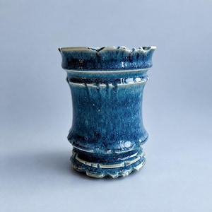 Puede incluir: Un jarrón de cerámica con un esmalte azul profundo, texturas moteadas y reflejos en azul claro. El recipiente presenta un borde acampanado, un cuerpo curvo y una base escalonada con crestas marcadas, mostrando un acabado brillante en toda la pieza.
