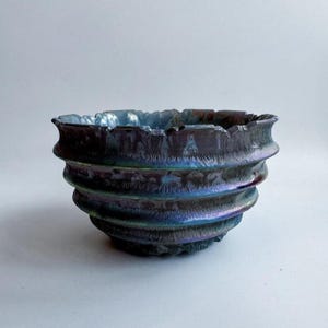 May include: A handcrafted ceramic bowl with a unique iridescent finish. The bowl features a dark, textured exterior with a rippled, layered design. The interior has a light blue hue. The bowl's dimensions are approximately 6 inches in diameter and 3 inches in height.