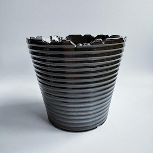 Op de afbeelding: Een donkerbruine, geribbelde keramische plantenbak met een unieke, gekartelde rand. De plantenbak heeft een glanzende afwerking en een cilindrische vorm, geschikt voor binnen of buiten. Het ontwerp heeft horizontale ribbels.