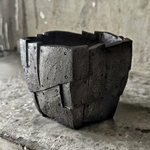 Puede incluir: Un cuenco de cerámica gris oscuro y texturizado con un diseño único similar a un tejido. El exterior del cuenco presenta una serie de segmentos rectangulares superpuestos, cada uno asegurado con pequeños remaches oscuros. El interior es liso y oscuro.