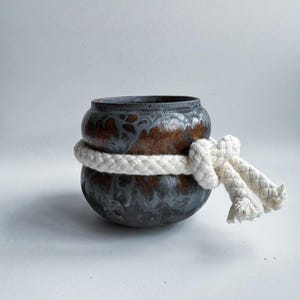 May include: A ceramic pot with a dark, mottled glaze in shades of brown and gray, encircled by a thick, white rope tied in a decorative knot. The pot has a rounded shape and a wide opening, suitable for decorative use.