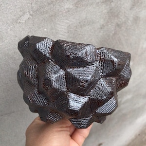 Può includere: Un esemplare di minerale marrone scuro e strutturato con una complessa struttura cristallina geometrica. La superficie riflette la luce, evidenziando gli intricati motivi e le forme. Il minerale è tenuto in una mano, mostrando le sue dimensioni e la sua forma.