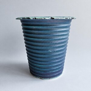 May include: A tapered ceramic vase featuring a deep blue and teal gradient finish. The exterior surface is defined by horizontal, ribbed ridges that wrap around the body, while the top rim has a textured, uneven edge with a light green glaze.
