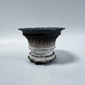 May include: A small, handcrafted ceramic pot with a unique design. The pot features a dark gray rim and a body that transitions from white to speckled brown. The base has a textured, rounded shape, perfect for small plants or decorative use.