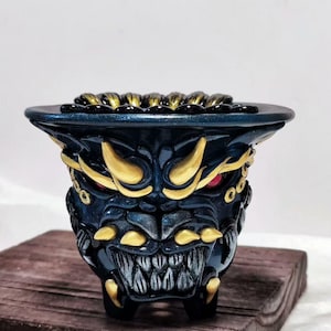 May include: A dark blue ceramic planter with a fierce, stylized demon face design. The planter features gold accents on the horns, teeth, and other details, with red eyes. The planter is on a wooden surface.