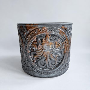Puede incluir: Una maceta cilíndrica con un relieve de un pulpo centrado dentro de un borde circular. La superficie tiene un acabado en gris oscuro y bronce desgastado con intrincados patrones geométricos que rodean el motivo central del pulpo.