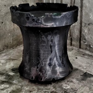 May include: A cylindrical black vessel with a flared top and bottom, featuring a rough, uneven, and textured surface. The rim has a jagged, notched edge, and the dark finish displays subtle variations in tone across the exterior body.