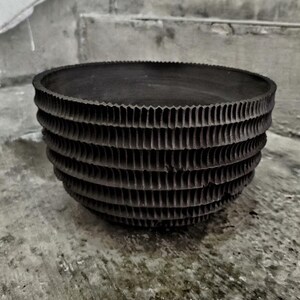 May include: A dark grey, round decorative bowl with a textured, layered design. The bowl features a series of horizontal, corrugated bands creating a unique, geometric pattern. The bowl is made of a solid material and is suitable for home decor.