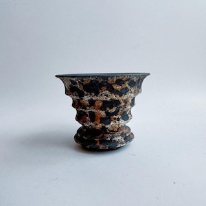Puede incluir: Un pequeño jarrón decorativo de cerámica con una superficie texturizada. El jarrón presenta un borde y una base negros, con un cuerpo adornado con un patrón moteado marrón y negro. El jarrón tiene una parte superior acampanada y una base de pedestal.