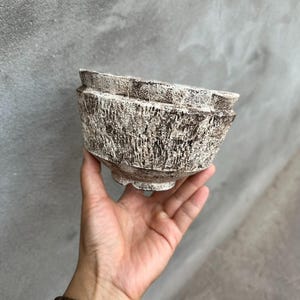 Op de afbeelding: Een getextureerde, gebroken witte keramische kom met een ruwe, gebeeldhouwde uitstraling. De kom heeft een brede, licht uitlopende rand en staat op een kleine, afgeronde basis. De kom wordt in een hand gehouden tegen een grijze achtergrond.