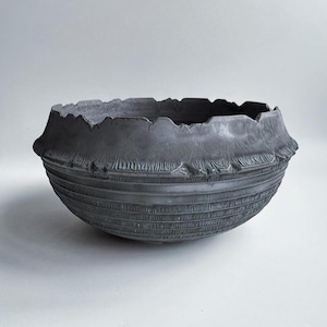 Puede incluir: Un cuenco gris oscuro texturizado con un diseño orgánico único. El cuenco presenta un borde áspero e irregular y patrones lineales horizontales alrededor del cuerpo. La estética general es rústica y moderna, adecuada para la decoración del hogar.