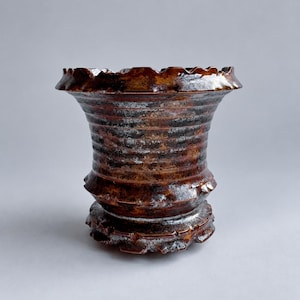 May include: A ceramic vase with a flared, scalloped rim and a wide, ribbed body. The surface features a mottled glaze in shades of deep brown, copper, and metallic silver, with a textured, uneven finish throughout the entire structure.