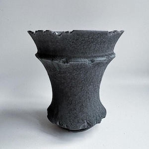 Puede incluir: Un jarrón de cerámica gris oscuro y texturizado con un borde acampanado y una superficie texturizada. El jarrón tiene una forma única y orgánica con una base ancha y una sección media más estrecha. El borde tiene un borde irregular y dentado.
