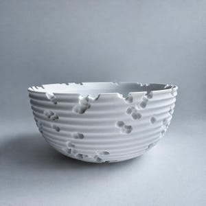May include: A white ceramic bowl featuring a series of horizontal ridges along its exterior surface. The bowl is punctuated by numerous irregular, crater-like indentations of varying sizes that break up the uniform ribbed pattern.