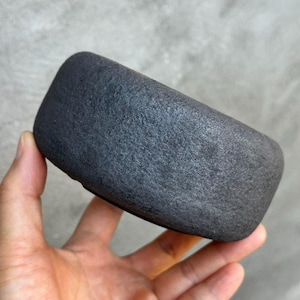 May include: A small, circular, dark charcoal-gray bowl with a rough, textured exterior surface. The object has a thick, rounded rim and a deep, open interior, showcasing a matte finish that appears porous and stone-like in its overall appearance.