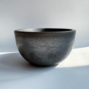 May include: A dark gray ceramic bowl with a speckled texture. The bowl has a rounded shape and a slightly flared rim. The interior is dark, and the exterior has a mottled appearance. The bowl is suitable for serving food or as a decorative piece.