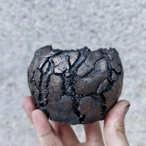 Puede incluir: Una pequeña maceta redonda de cerámica de color oscuro con una superficie texturizada y agrietada. La maceta se sostiene en una mano y parece no estar esmaltada. El color es una mezcla de marrón oscuro y negro.