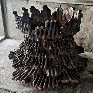 Puede incluir: Un jarrón decorativo texturizado de color bronce con un diseño orgánico único. El jarrón presenta una estructura calada en capas con una parte superior dentada en forma de corona. La estética general es rústica y artística.