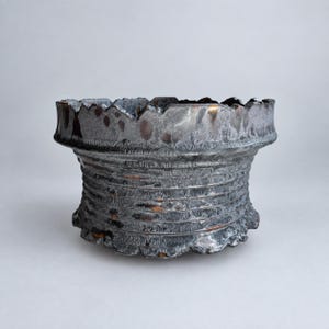 May include: A wide, cylindrical ceramic bowl with a dark, metallic, and iridescent glaze. The exterior features horizontal ridges and a textured, uneven surface. The rim is jagged and irregular, showing hints of copper and bronze tones.