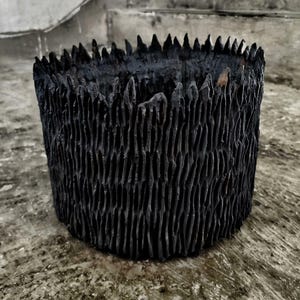 Peut inclure: Pot cylindrique en céramique noire avec un motif texturé à rainures verticales. Le bord présente des bords dentelés et pointus. Le pot est posé sur une surface en béton, mettant en valeur son design unique et artisanal. Idéal pour la décoration intérieure ou comme pot de fleurs.