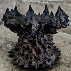 Könnte beinhalten: Dunkle, strukturierte Skulptur, die an eine Krone oder ein stacheliges Gefäß erinnert. Das Stück ist überwiegend schwarz mit braunen Akzenten, mit einer rauen, unebenen Oberfläche und spitzen Vorsprüngen. Auf neutralem Betonhintergrund.