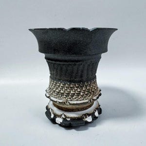 Puede incluir: Un jarrón de cerámica negra con borde festoneado. El jarrón tiene un diseño texturizado y estampado en tonos negros, blancos y marrones. La base tiene un diseño decorativo similar a una flor.