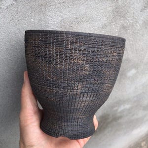 May include: A dark brown, textured ceramic bowl with a flared top and a narrow base. The bowl has a woven basket-like pattern. The bowl is held in a hand against a gray background.