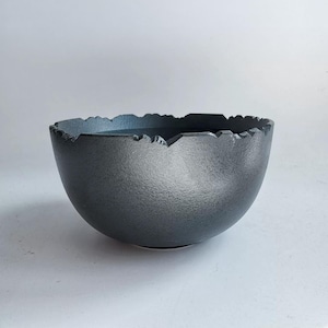 May include: A dark gray bowl with a textured, matte finish and a jagged, uneven rim. The bowl has a deep, rounded basin and a solid, heavy appearance, featuring a rough, stone-like surface texture throughout the exterior and interior.