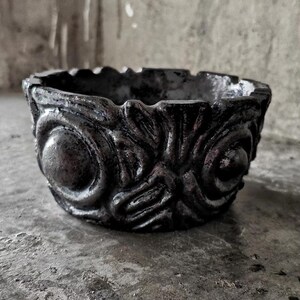 Puede incluir: Un cuenco circular de color gris oscuro que presenta un patrón de relieve texturizado en su exterior. El diseño consiste en motivos simétricos y arremolinados y elementos circulares prominentes que rodean toda la circunferencia.