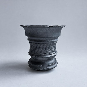 May include: A dark grey, textured ceramic vessel with a wide, flared rim and a tapered body. The surface features a prominent, raised basket-weave pattern around the centre, complemented by decorative, notched ridges at the base and midsection.