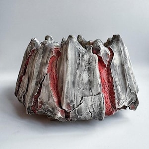 Puede incluir: Un cuenco de cerámica artesanal con un diseño abstracto y texturizado. El exterior presenta una combinación de gris y blanco con líneas verticales, mientras que el interior revela un vibrante tono rojo. La forma irregular del cuenco realza su atractivo artístico.