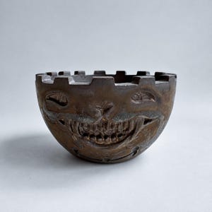 May include: A dark brown ceramic bowl with a crenellated rim and a sculpted face featuring narrow eyes and a wide, toothy grin. The surface has a metallic, weathered finish with visible texture and depth across the moulded facial features.