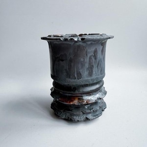 Puede incluir: Una maceta de cerámica gris oscuro con una superficie texturizada. La maceta tiene un borde acampanado y una base decorativa con un diseño en capas. El color es una mezcla de gris oscuro y marrón, con acentos blancos.