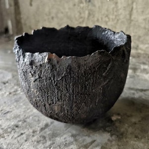 Puede incluir: Un cuenco de color carbón oscuro con una superficie rugosa y texturizada. El cuenco tiene un borde irregular y dentado y una base redondeada. La pieza de cerámica hecha a mano probablemente se utiliza con fines decorativos.