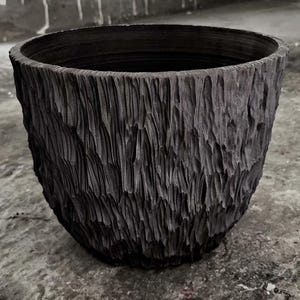 Puede incluir: Una maceta gris oscuro con una textura rugosa, con aspecto de madera tallada. El recipiente redondo con la parte superior abierta tiene una boca ancha y un exterior texturizado, adecuado para uso en interiores o exteriores. La maceta está hecha de un material duradero.