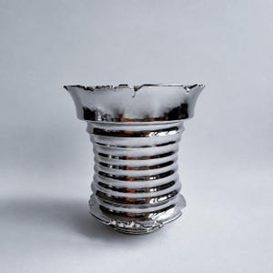 以下が含まれることがあります： 金属光沢のある反射性の銀色の花瓶。胴体には一連の水平な溝があり、縁は幅広く広がっており、不規則なスカラップ状の端が特徴です。表面は高度に研磨されており、光を鮮明に反射しています。
