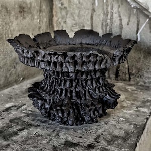 Puede incluir: Un cuenco decorativo negro, texturizado, con un diseño orgánico único. El cuenco tiene un borde ancho y con volantes y una base texturizada y en capas. El cuenco está hecho de un material oscuro y se asienta sobre una superficie de hormigón.