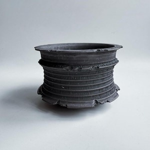 Könnte beinhalten: Ein dunkelgrauer, zylindrischer Keramiktopf mit einem strukturierten, gerippten Design. Der Topf hat einen breiten, flachen Rand und einen leicht ausgestellten Boden. Das Äußere weist horizontale Rippen und eine raue, unebene Textur auf.