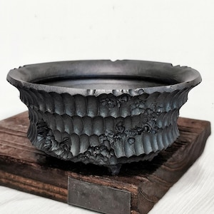 Puede incluir: Una maceta de cerámica gris oscuro texturizada con un diseño floral detallado. La maceta se asienta sobre una base de madera oscura. La maceta tiene una forma ovalada ancha y tres pequeños pies.