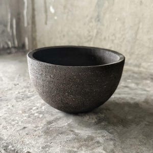 May include: A dark gray, semi-spherical bowl with a textured surface. The bowl has a wide opening and a rounded base. The interior is a deep black, contrasting with the exterior's lighter gray tones. The bowl is likely made of stone or a similar material.