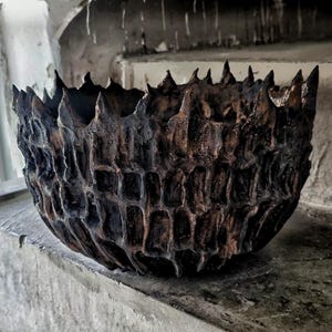 May include: A dark brown and black ceramic bowl with a highly textured, rugged exterior featuring deep vertical grooves and a jagged, spiked rim. The surface has a matte, weathered appearance with irregular indentations throughout the vessel.
