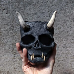 Könnte beinhalten: Ein schwarzer, schädelartiger Pflanzentopf mit weißen Hörnern und Fangzähnen. Der Topf hat eine strukturierte Oberfläche und wird in einer Hand gehalten. Der Schädel hat leere Augenhöhlen und eine detaillierte Gesichtsstruktur.