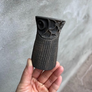 Puede incluir: Objeto de madera tallada de color marrón oscuro con un cuerpo cilíndrico texturizado y una parte superior detallada. La parte superior presenta un diseño en forma de ojo y tallas florales. El objeto se sostiene en una mano sobre un fondo gris.