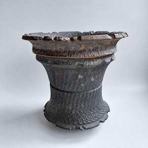May include: A dark grey ceramic vessel with a wide, flared rim and a textured, rippled surface. The upper edge features a metallic bronze-toned finish that transitions into a deep charcoal body with a repeating wave-like pattern.